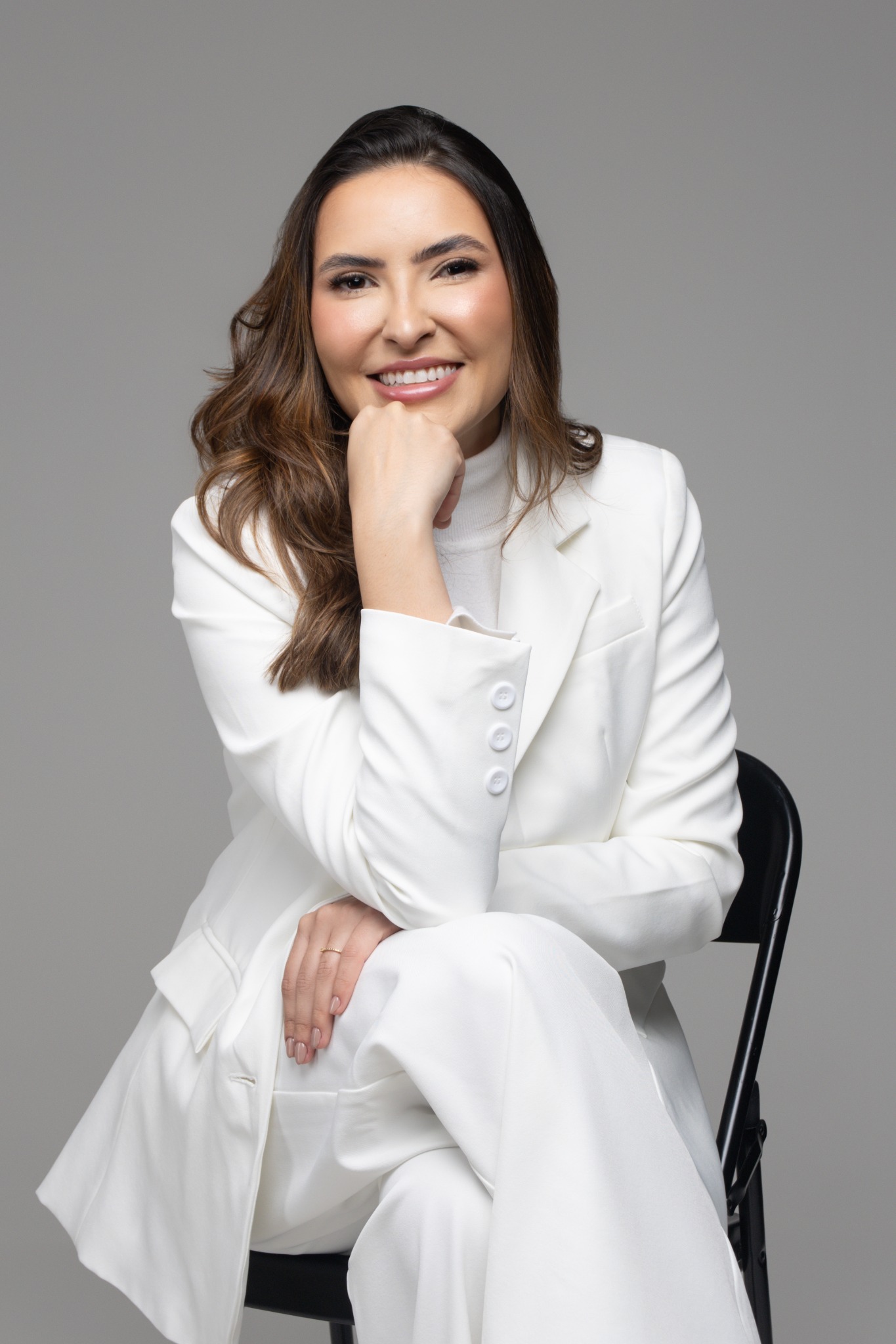 Dra. Kathellyn sentada, sorrindo, com a mão apoiada no queixo e conjunto branco